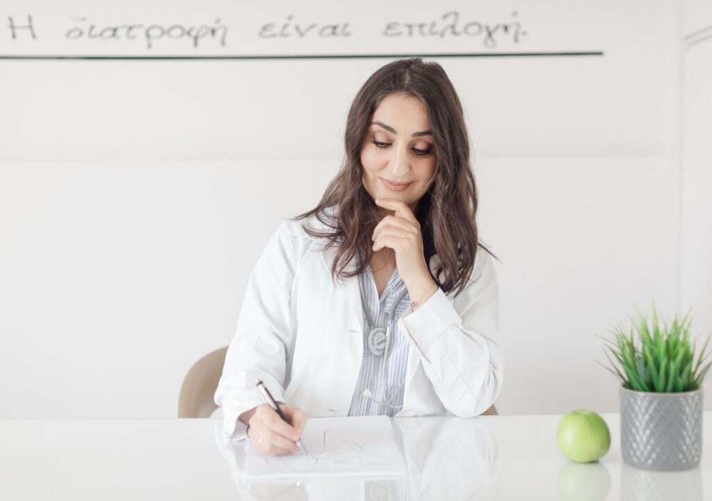 A female nutritionist in a lab coat writing notes with an apple and plant on the desk.