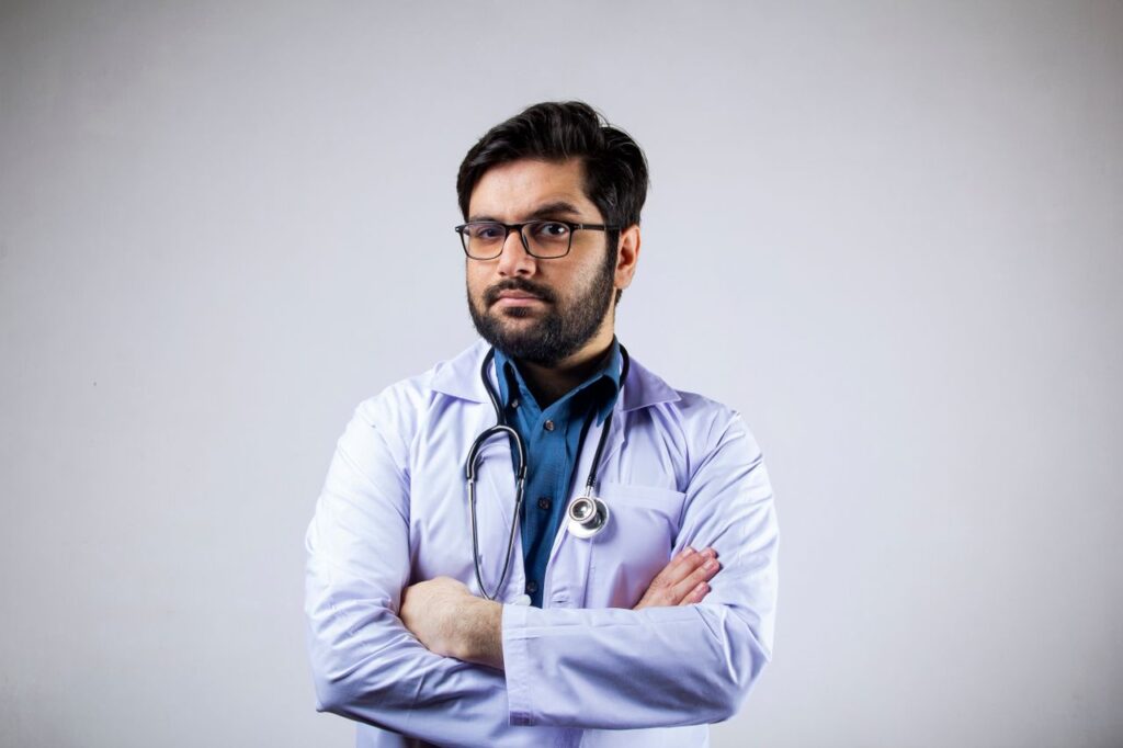 About 1 Portrait of a serious male doctor in a white coat with arms crossed, wearing glasses and a stethoscope.